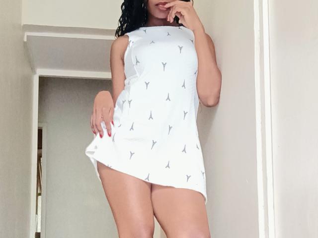 NalediJones stands in a white dress with small black airplane patterns, showing her ass and thighs, with one hand near her mouth, black curly hair, red nails.