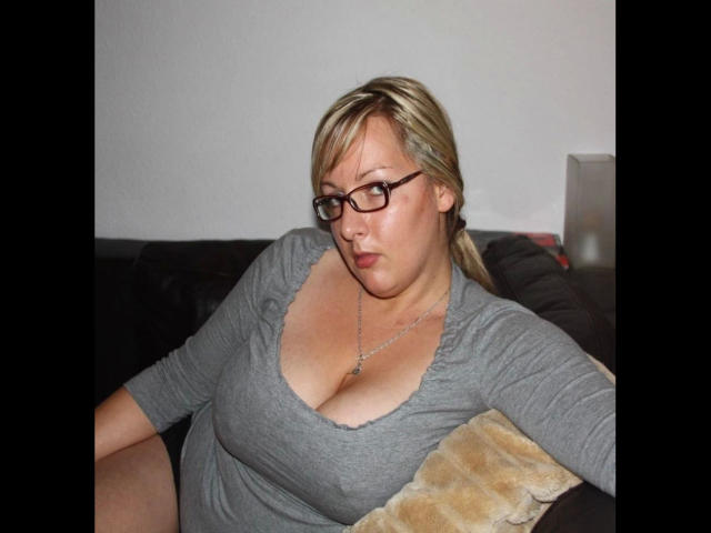 Mara-80F, a plus-sized woman with blonde hair and glasses, sits on a dark couch wearing a grey, low-cut top that reveals her big, round, and slightly saggy tits.