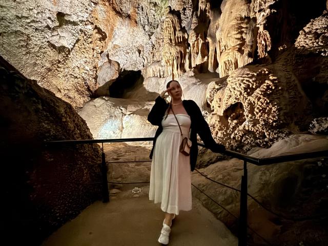 MonaPlus with big ass, standing in a rocky cave, wearing a white dress, black jacket, white socks, and a brown crossbody bag, holding a phone to her ear.