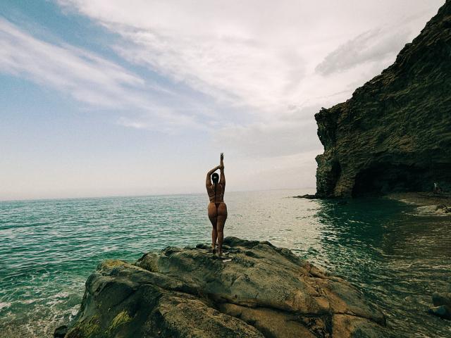 MonaPlus stands on rocky shore, arms in yoga pose, back to camera, showing a round ass, with ocean and cliff in background. Clear blue sky, white clouds.