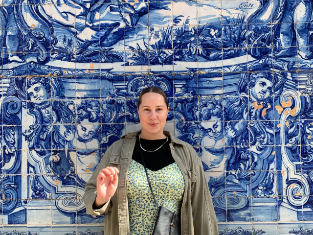 MonaPlus stands in front of blue ceramic tiles, wearing a green jacket over a floral top, with a black necklace and a silver cross, holding up her right hand.