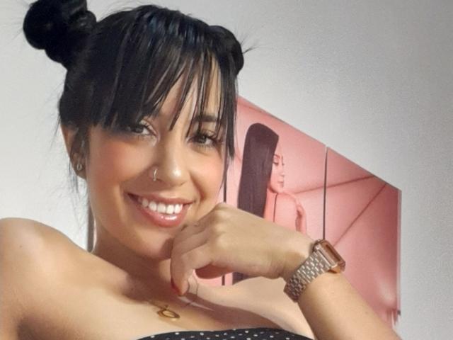AnalisseJade with black double buns, smiling, showing her shiny teeth, wearing a strapless black top, nose ring, and gold watch, standing in front of a pink portrait.