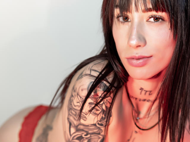 MiaNapoles with dark hair, tattoos on her arm and neck, wearing red lace panties, has brown eyes and pink lips, looking at the camera with a slight smile.