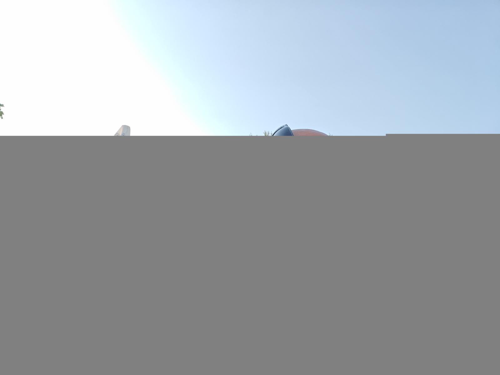 Het hoofd van CandyHotCouple69, met een blauwe hoed en een rode hoed die gedeeltelijk zichtbaar is boven een grijze muur, tegen een helderblauwe lucht..