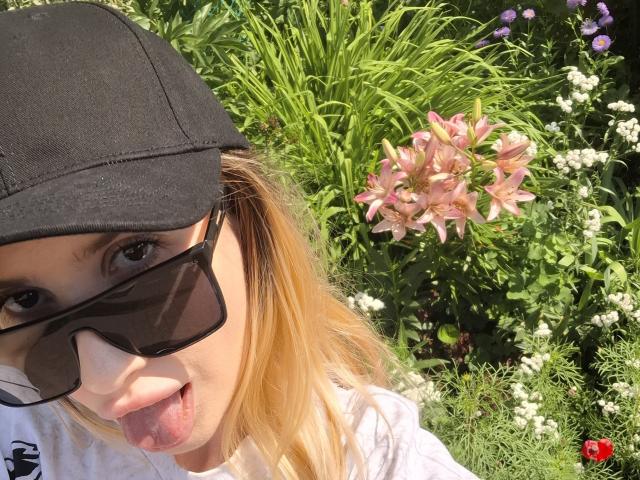 ElzaKomx with blonde hair, black cap, sunglasses, and tongue out, wearing a white shirt, in a sunny garden with pink lilies and green foliage.