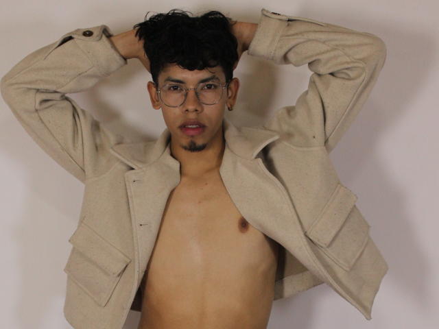 LianStonne, shirtless, beige jacket, hands in messy black hair, round glasses, small goatee, light brown skin, visible nipple, neutral expression, white background.