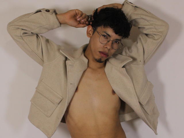 LianStonne, shirtless, wearing beige jacket, arms raised, glasses, short curly black hair, light brown skin, small goatee, looking at camera, chest visible, slight nipple shadow.
