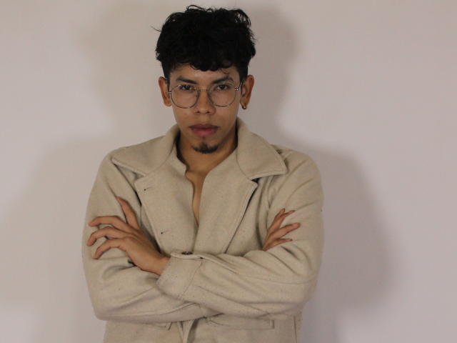LianStonne with curly black hair, wearing round glasses, a beige coat with arms crossed, and a small goatee, standing against a plain white wall.