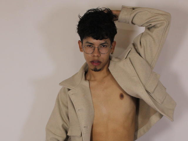LianStonne, shirtless, with beige jacket, hand in messy black hair, wearing glasses, small brown nipples, slightly open mouth, light skin, dark curly hair, subtle facial stubble.
