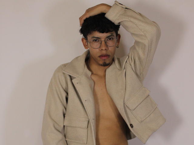 LianStonne with curly black hair, glasses, and a beige jacket, shirtless, hand in hair, looking at camera, beige wall background, subtle beard, small earrings.