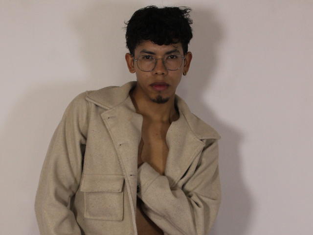 LianStonne with curly black hair, glasses, and gold hoop earring, wearing an open beige coat, exposing a small chest, looking serious with a slight open mouth.