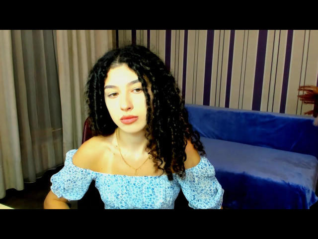ArianaFoxx, avec ses cheveux noirs bouclés, sa peau claire et ses lèvres rouges, portant une robe bleue à fleurs avec une épaule dénudée, assise devant un canapé en velours bleu..