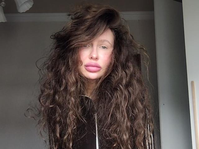 AnnPat with long, messy brown hair covering her eyes, pink lips, and a nose piercing, wearing a black shirt, standing in a room with white walls.