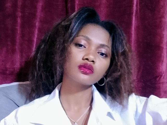 Cassiy has dark, wavy hair, purple lipstick, hoop earrings, and a white shirt, looking serious with a slight pout. Her ass is not visible. Red curtain background.