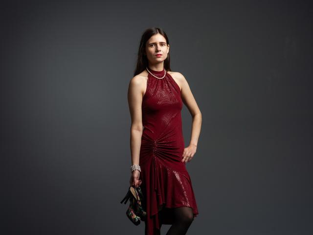 MartaLeee stands in a dark room, wearing a red, sleeveless dress with a ruched skirt, holding floral heels in her right hand, with a serious expression.