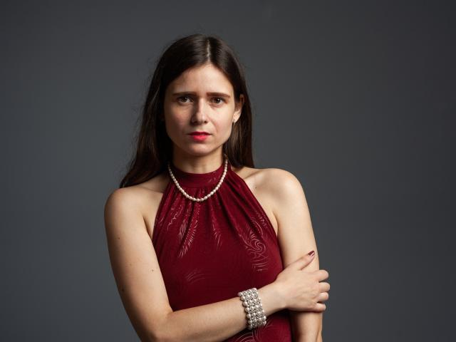 MartaLeee with dark brown hair, red lipstick, wearing a red halter dress, pearl necklace, and pearl bracelet, arms crossed, looking serious. Boobs are small, ass is not visible.