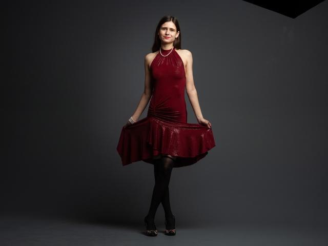 MartaLeee has pale skin, long brown hair, and small tits. She's wearing a red dress, black stockings, and black heels, holding the dress out with a cute smile.