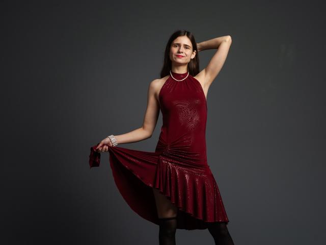 MartaLeee, with long brown hair, wearing a red halter dress, holding it out, smiling, arm raised, black thigh-highs, silver bracelet, dark gray background, standing confidently.