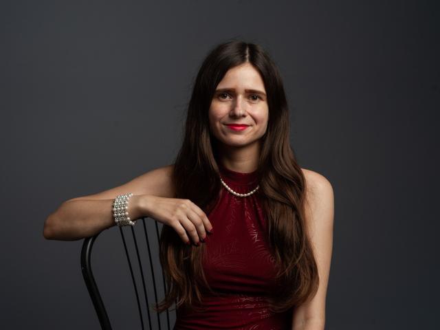 MartaLeee with long brown hair, red dress, pearl necklace, and bracelet, smiling, resting arm on black chair, dark background. Boobs are not visible. She has red lipstick.