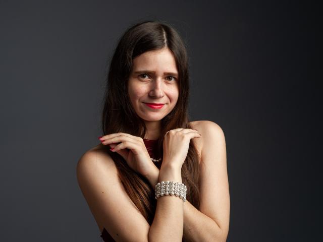 MartaLeee with long brown hair, red lipstick, and a sleeveless maroon top, smiling, hands near neck, wearing a silver bracelet, dark background. Boobs not visible.