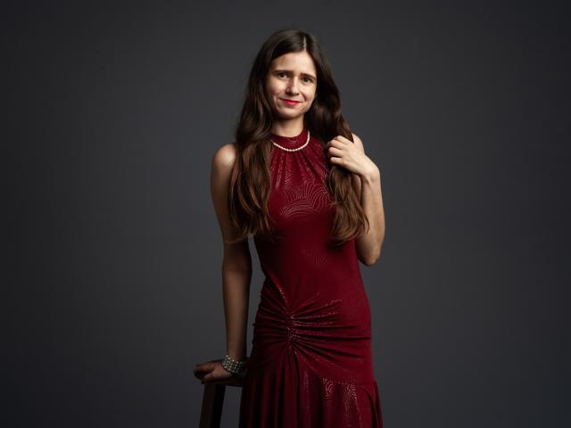 MartaLeee with long brown hair, red dress, and pearl necklace, standing, hand on chair, smiling, red lipstick, dark background. Boobs are covered by dress, ass not visible.