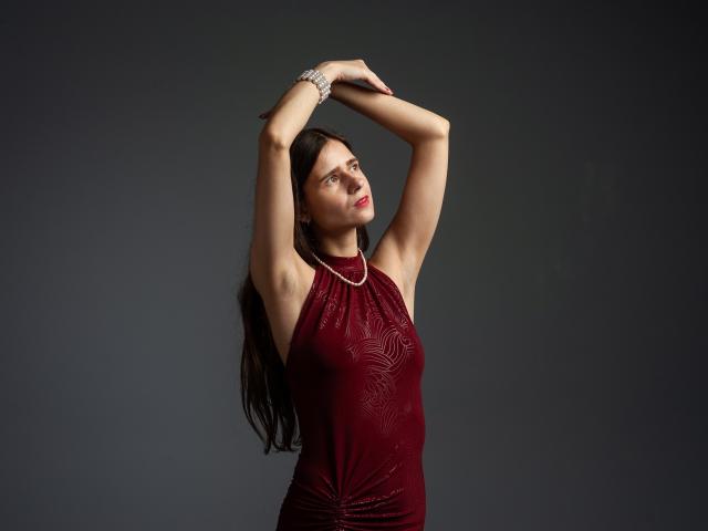 MartaLeee with arms raised, wearing a sleeveless red dress with a leaf pattern, silver bracelet, and necklace, looking slightly up with red lipstick. Dark background.