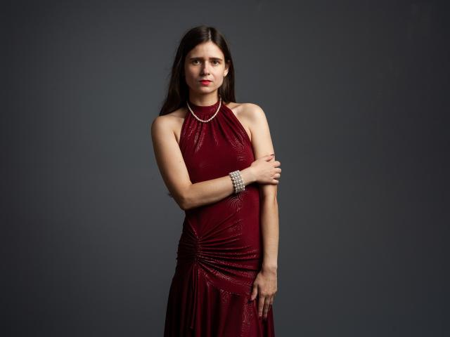 MartaLeee with long brown hair, wearing a shiny red dress, covering her small tits with her arm, silver bracelet, and necklace, standing against a dark gray background.