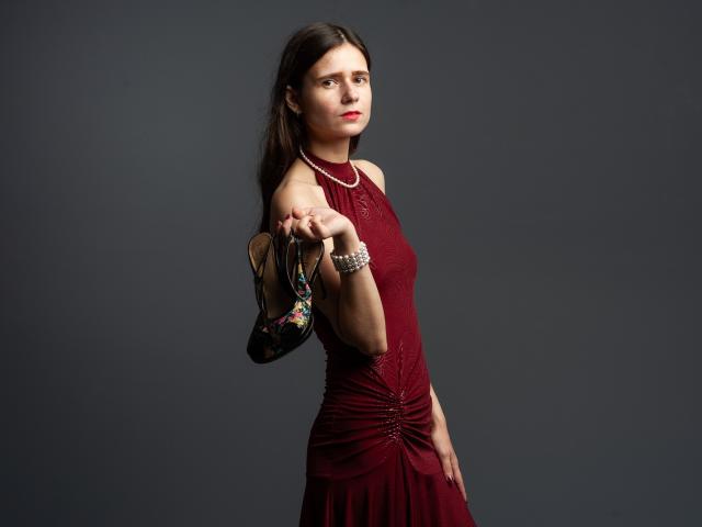 MartaLeee with long brown hair, red dress, holding black floral heels in right hand, standing sideways, wearing pearl necklace and bracelet, gray background.