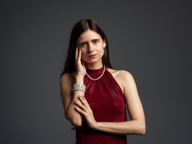MartaLeee with long dark hair, wearing a red halter dress, pearl necklace, and sparkly bracelet, touching her face, looking serious, with red lipstick and bare shoulders.