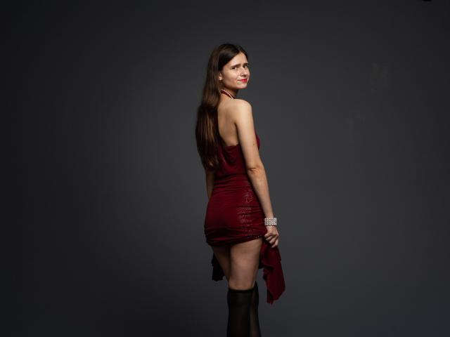 MartaLeee stands with back to camera, wearing a red dress, holding it up to expose her bare ass, in black thigh-highs, with long brown hair and red lipstick.