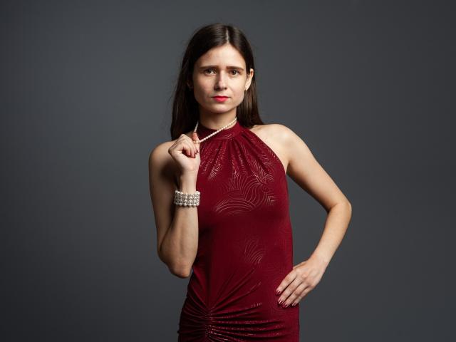 MartaLeee has long brown hair, wearing a red, halter-neck dress with subtle patterns, and a pearl bracelet. She's holding a necklace, with her hand on her hip.
