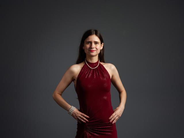 MartaLeee with long brown hair, red dress, and sparkly bracelet, standing with hands on hips, smiling, showing her tits and ass in a dark studio.