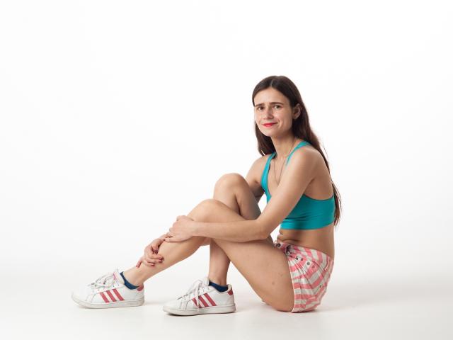 MartaLeee sitting with legs crossed, wearing a teal sports bra, pink plaid shorts, white Adidas sneakers with red stripes, and a slight smile. Her tits are small.