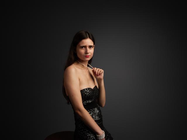 MartaLeee with long brown hair, wearing a black lace strapless dress, holding a toothbrush near her red lips, and a silver bracelet on her right wrist.