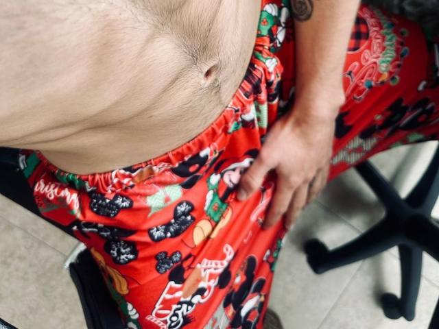 JakeMagnus's bare, hairy chest with a visible navel, wearing red Christmas pajama pants featuring black and white Mickey Mouse prints, sitting on a chair with one hand on his pants.