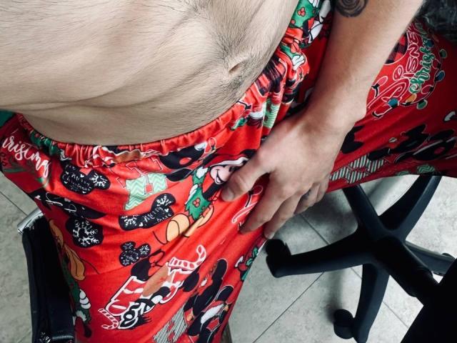 JakeMagnus's hairy belly, wearing red Christmas pajama pants with Mickey Mouse designs, sitting on a black office chair, hand resting on his crotch, showing a tattoo on his arm.