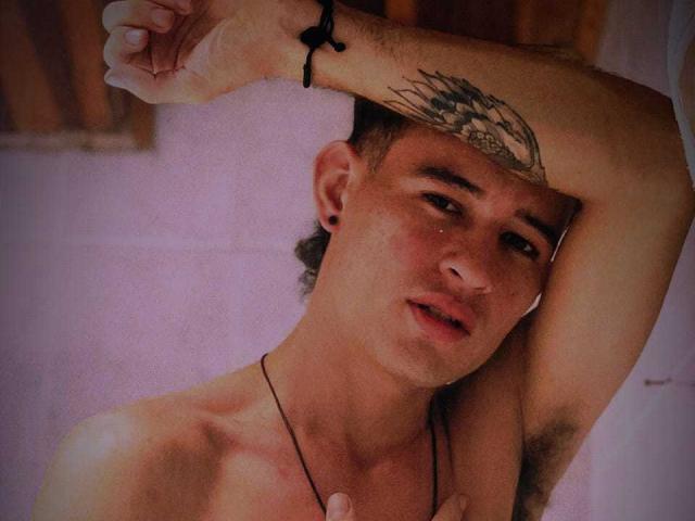 PosionAngel, shirtless, arm raised, tattoo on forehead, short brown hair, black ear hole, black necklace, slight blush, relaxed, indoor, soft light, slight smile, casual, intimate.
