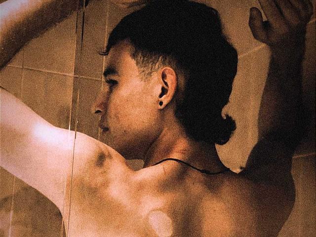A topless PosionAngel with a butt, short dark hair in a bun, black ear plugs, and a necklace, raises arms against a glass wall in a dimly lit shower.