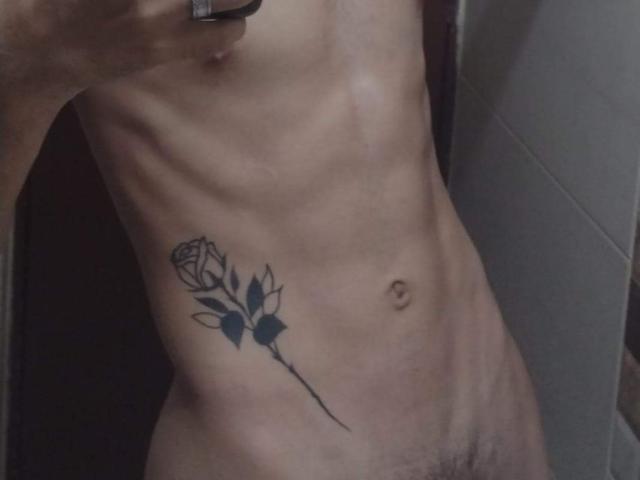 Slim, shirtless PosionAngel with a black rose tattoo on their left side, exposing their flat stomach and a small, visible navel. Their right hand holds a phone.