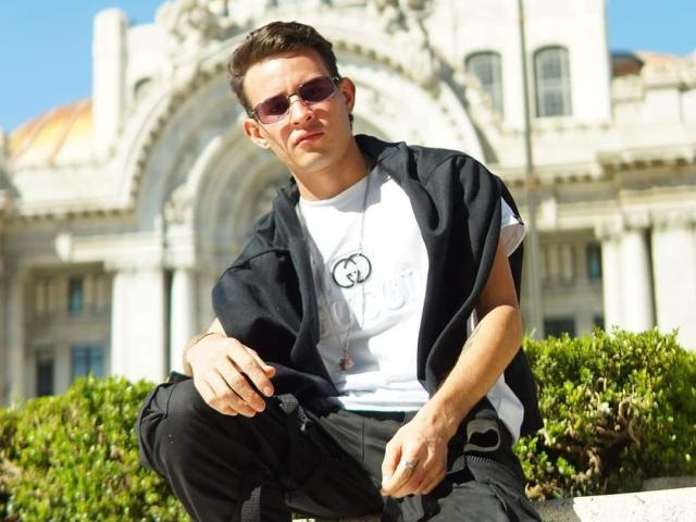PosionAngel with short brown hair, sunglasses, white tee, black jacket, black pants, and necklace, squatting, green bushes, white building with arches in background, sunny, smiling.