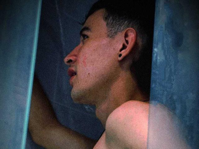 A shirtless PosionAngel with short dark hair and black ear studs peeks through a blue-tinted glass panel, showing a slightly flushed face and a focused expression.