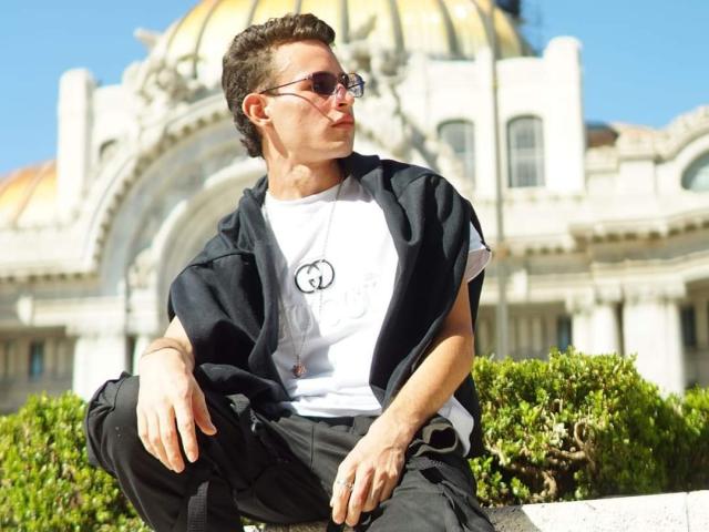 PosionAngel with short brown hair, sunglasses, white tee, black shirt draped over shoulders, black pants, sitting on a stone ledge, looking right, in front of a white building with a dome.