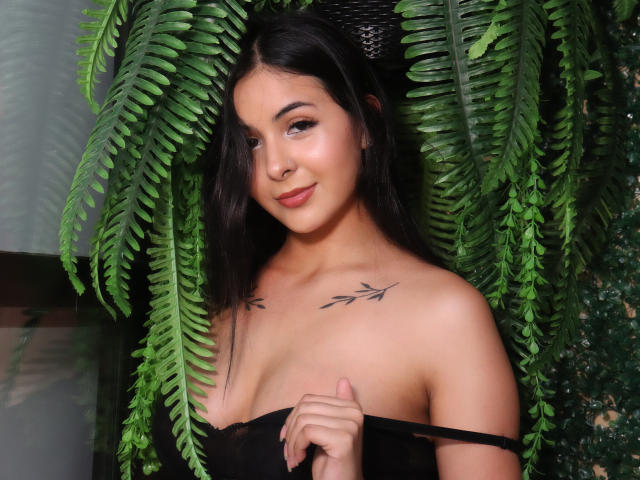 GeorginaGb with dark hair, brown eyes, and tattoos on her chest, holding a black strap, standing in front of green ferns, smiling softly. Boobs visible, no clothes on top.