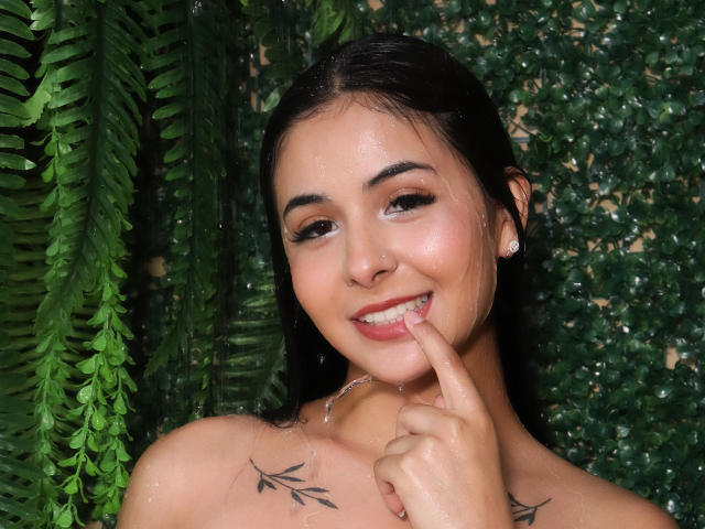 GeorginaGb, wet, smiling, black hair, tattoos on shoulders, finger in mouth, green leafy background, water droplets on skin, earrings, looking up, attractive, cute, sexy.
