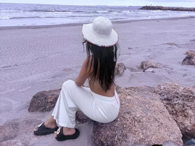 AlisonKweel sitzt auf Felsen, trägt weiße Hosen, ist obenhin nackt, hat langes, dunkles Haar, einen weißen Hut und schwarze Flip-Flops und blickt auf das Meer. Ihr Hintern ist leicht sichtbar..