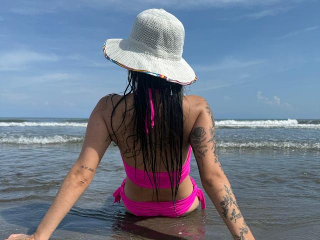 AlisonKweel with wet black hair, wearing a white sun hat and pink bikini, sitting in shallow ocean water, showing tattoos on her arms and back, with a blue sky.
