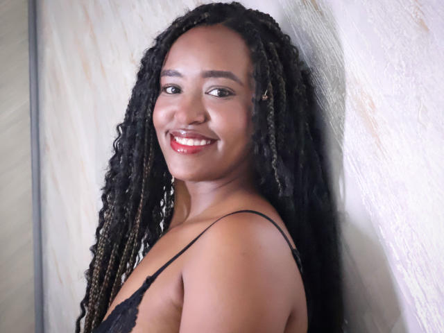 NaomiReyes with dark brown skin, long black braided hair, wearing a black lace top, smiling with red lipstick, showing her teeth, standing against a white textured wall.