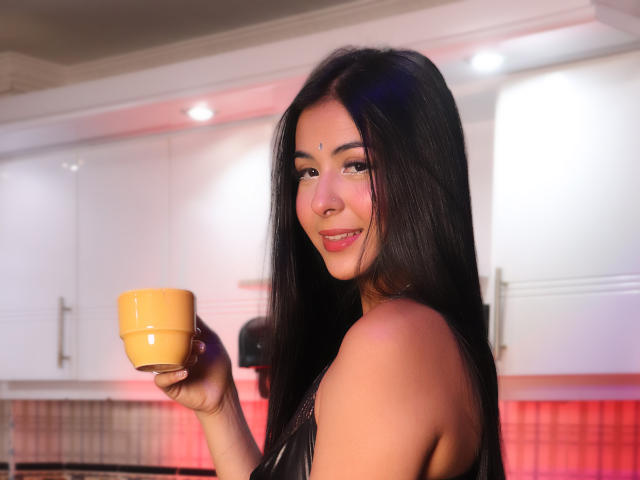 GeorginaGb with long black hair, holding a yellow cup, smiling at the camera, showing her nose ring, in a kitchen with white cabinets and red lighting.
