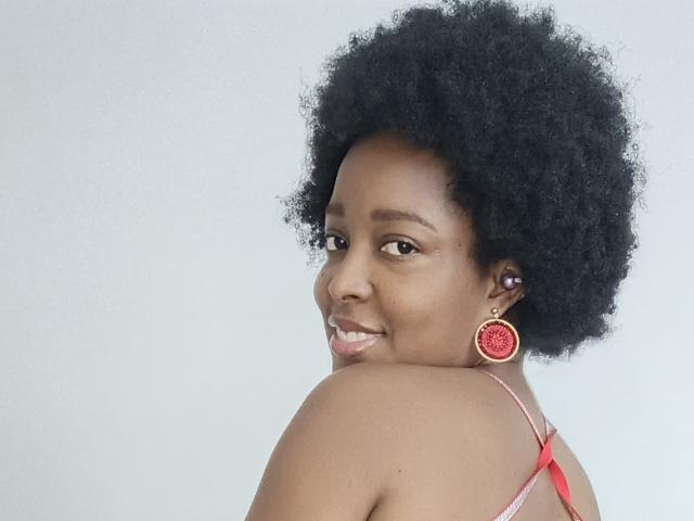 ChelseyeBonny with a big afro, smiling at the camera, wearing red earrings and a pink strappy top, showing her shoulder and upper back.