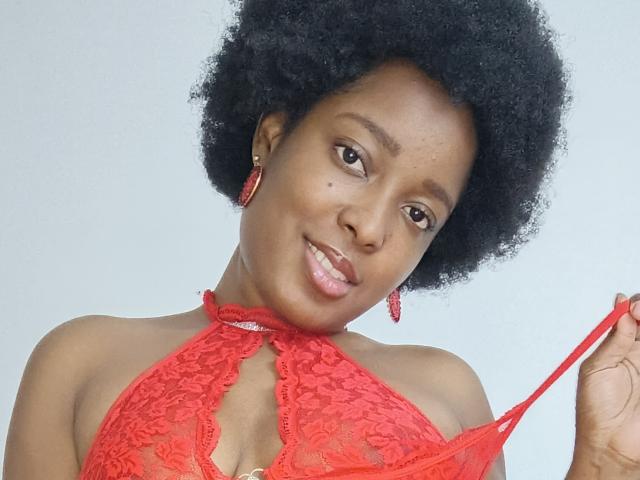 ChelseyeBonny with a big afro, wearing a red lace halter top, pulling down the strap, showing her tits, smiling, wearing red earrings, against a light background.